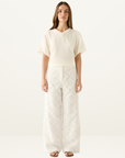 Model wearing white wide-leg pants featuring intricate eyelet embroidery and a matching cream V-neck t-shirt.