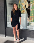 Woman wearing a black cotton mini tee dress with a cinched waist, pleated side detail, and silver rod accent.