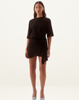 Model wearing a black cotton mini tee dress with elbow sleeves, cinched waist, and pleated asymmetric skirt with silver rod.