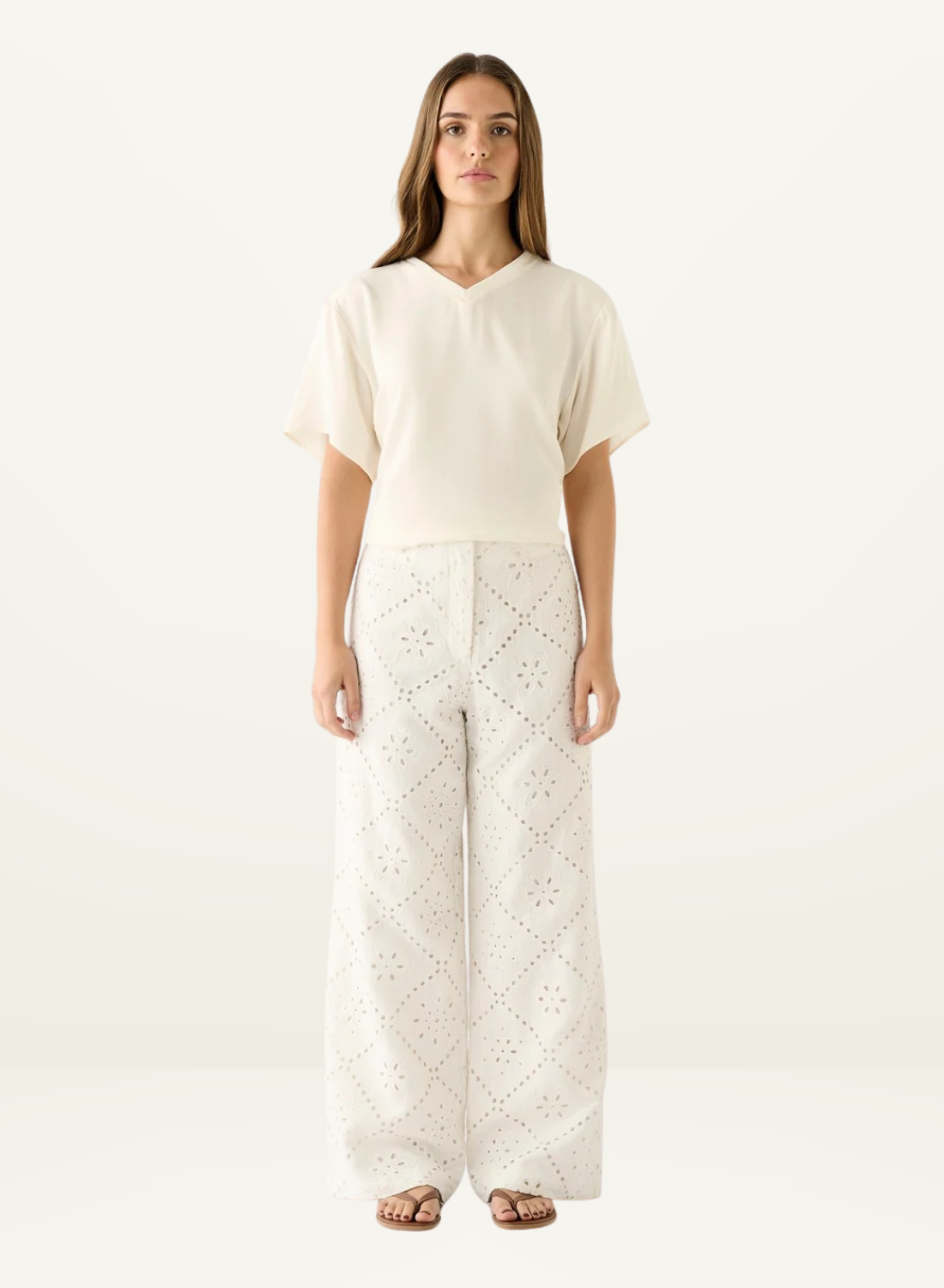 Model wearing white wide-leg pants featuring intricate eyelet embroidery and a matching cream V-neck t-shirt.